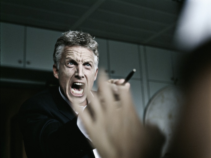 A man in a suit appears to be speaking passionately or engaging in stress management, holding a pen and gesturing with his hand.