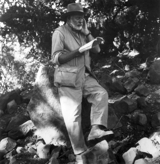 Ernest Hemingway is reading book & siting on rock.