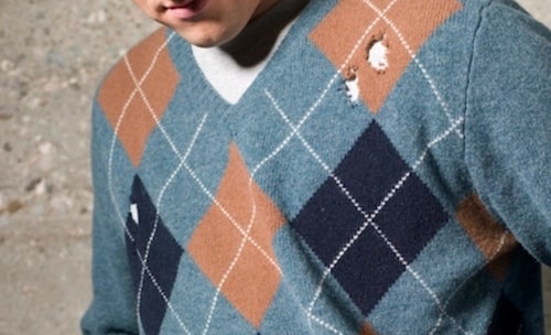 A young man in a blue sweater leaning against a wall.