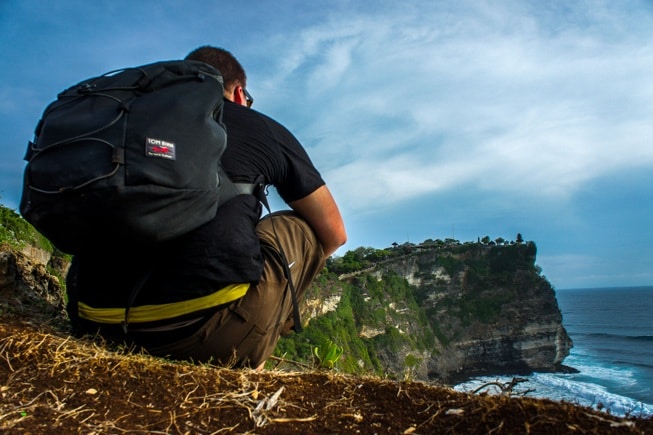 A man with a backpack sitting on a cliff overlooking the ocean, contemplating the vast world before him.